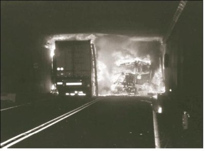View through a tunnel where an explosion has taken place. The rear view of a truck is shown in the left lane while remnants of other vehicles and debris are in flames in the right. The view beyond is obscured by flames and smoke.