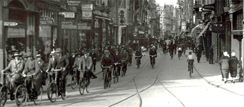 Figure 4: Amsterdam during peakhour( 1934) - Description: A black and white picture of cyclists in the streets of Amsterdam dressed in suits and top hats in 1934.