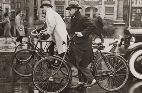Figure 5: Amsterdam during peakhour( late 1930s) - Description: A black and white closeup of two men in peacoats and top hats riding their bike. 