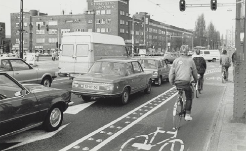 Figure 21 Temporary bilke lane in Berlagebrug - Description: This picture shows two lines of traffic stopped at a red light with individuals riding in a bike lane made on the right of the two lanes of traffic.
