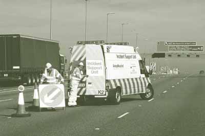Pictured is a van with an expanded roofline, painted with bold diagonal stripes and the words "Incident Support Unit" and "AmeyMouchel," from which two men in reflective gear are shown setting up reflective traffic cones and arrow signage.
