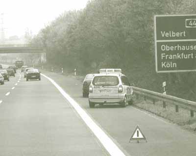 Titled, "ADAC on German motorway," this photograph depicts a German automobile club van stopped behind a disabled vehicle in the shoulder of a highway, with a triangular marker set up behind it.