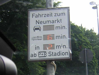 Photo of travel time comparison display on Cologne arterial showing travel time in minutes by car and by public transport and how many minutes until the next tram arrives.