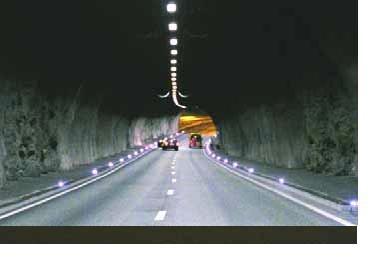 Photo looking through a tunnel with lights running along the top center of the tunnel as well as along the outer edges of the roadway on both sides.