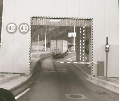 Photograph of a truck taken through the passageway of the Truck thermal detector. The truck, seen from behind, is making it's way to the tunnel entrance having already passed through the detector. A lift gate is in the down position with a traffic signal mounted on a post to the right of the passageway. The passageway door perimeter is marked with a border of diagonal contrasting stripes.